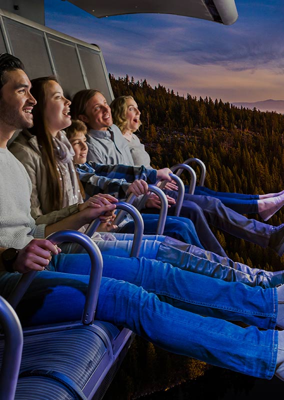 A group of people sit on a flight ride over a scene of forests and a lake.