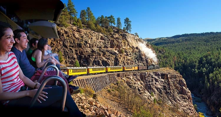Riders on the flight ride enjoy the ride as a train rides on a cliff side.